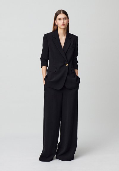 Blazer negro ajustado con un solo botón dorado, combinado con pantalones de pierna ancha negros. La tela parece lisa, con un ajuste estructurado.