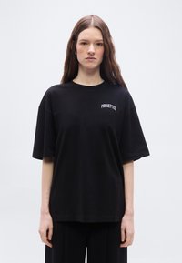 Νεαρή γυναίκα με μακριά καστανά μαλλιά, που φοράει ένα υπερμεγέθες μαύρο T-shirt με την επιγραφή «PIROUETTES» στο στήθος, στέκεται μπροστά σε απλό φόντο.