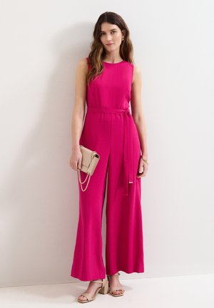 Femme portant une combinaison rose vif sans manches à jambes larges, tenant une pochette beige, portant des sandales beiges à talons blocs, debout contre un mur blanc.
