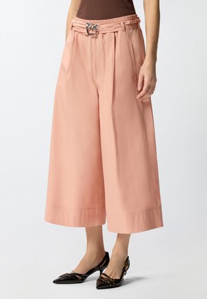 Donna che indossa pantaloni rosa a gamba larga a vita alta corti, con una cintura decorativa e décolleté slingback nere con punta affilata.