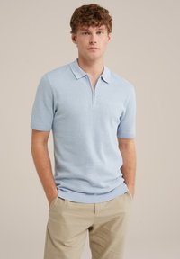 Tricou polo cu mânecă scurtă de culoare bleu deschis, cu guler cu fermoar, dintr-un material tricotat texturat și cu margini ribbate. Asortat cu pantaloni bej.