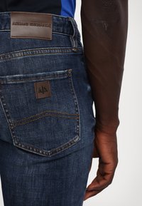 Tumši zili džinsi ar pieguļošu formu. Aprīkoti ar ādas birku ar "Armani Exchange" logotipu un mazu kabatas detaļu.