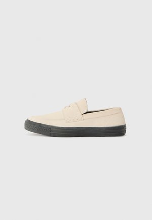 Beige slip-on cipő sima velúr felsőrészel és fekete gumitalppal. Kiemelkedő részlet a cipő orrán, valamint alacsony profilú dizájn.