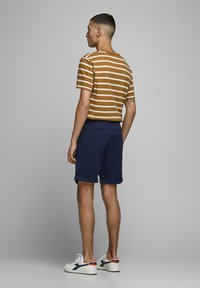 T-shirt rayé marron et blanc associé à un short bleu marine. Le modèle porte des baskets blanches avec des accents bleu marine. Textures lisses tout au long.