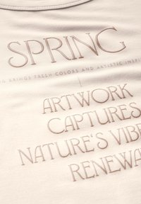 T-shirt beige clair avec texte en relief et en relief : "LES ŒUVRES D'ART DU PRINTEMPS CAPTURER LES VIBRATIONS DE LA NATURE RENOUVELLEMENT." Texture de tissu douce et lisse.