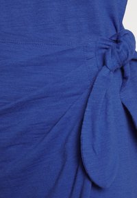 Modrá tunika vyrobená z lehkého materiálu, s uzlovým detailem na jedné straně a mírně texturovaným povrchem.