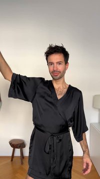 Črno satenasto haljo z ovijalnim dizajnom, V-izrezom, kratkimi rokavi in pasom, ki se veže. Gladek tekstura in minimalni poudarki.