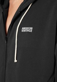 Juodas užtrauktukas su reguliuojamu raiščiu. Yra "AMERICAN VINTAGE" logotipas baltai kairėje krūtinės pusėje. Minkšta, tekstūruota medžiaga.