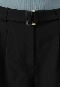 Czarne, dopasowane spodnie wykonane z gładkiego materiału, z zakładkami z przodu i srebrną metalową klamrą na pasku.
