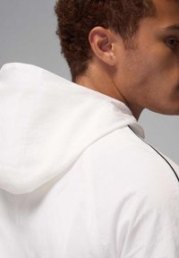 Chaqueta blanca con capucha de tela texturizada, con un detalle negro a lo largo de la costura del hombro y una capucha suave y caída.