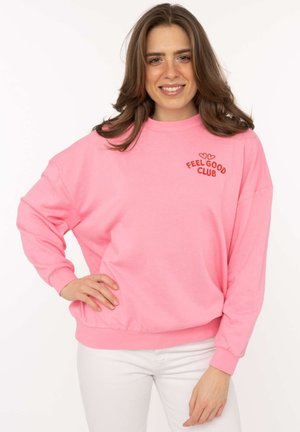 Junge Frau trägt ein pinkfarbenes Sweatshirt mit dem Schriftzug "Feel Good Club", steht mit einer Hand in der Hüfte und lächelt vor einem einfachen weißen Hintergrund.