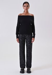Maglione nero con spalle scoperte abbinato a pantaloni neri glitterati. L'outfit presenta dettagli lucidi e una vestibilità rilassata, completato da scarpe nere chunky.