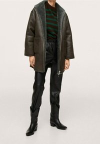 Bruine leren oversized jas met een groene nepbontkraag, gecombineerd met zwarte leren broeken en zwarte cowboylaarzen. Gestreepte trui eronder.