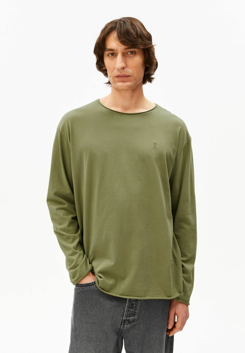 Man in olijfgroene lange mouwen en donkergrijze jeans, staand met één hand in de zak tegen een effen witte achtergrond.