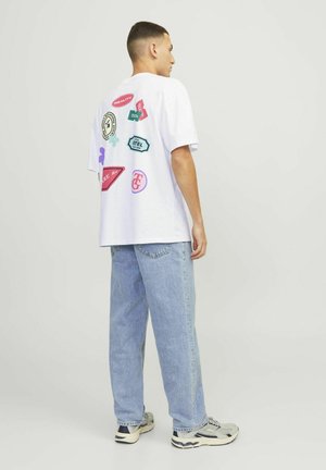 White graphic t-shirt with various colorful patches, paired with light blue jeans. The shirt features a loose fit and round neckline.