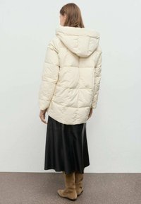 Chaqueta de plumas crema con capucha, que presenta un diseño acolchado. Llevada sobre una falda negra larga y botas marrón claro hasta la rodilla.