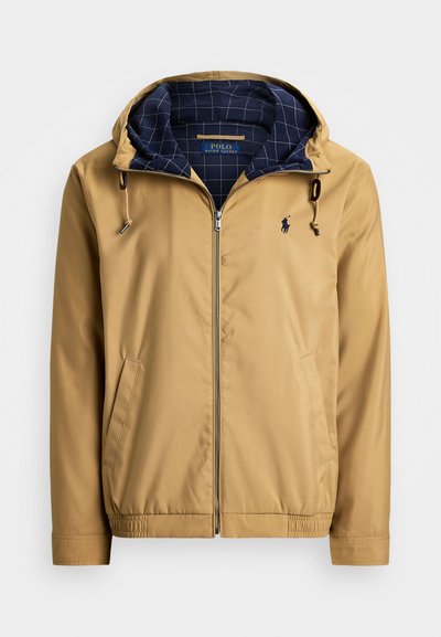 Beige zipjakke med justerbar hette. Har marineblå rutete fôr, sidelommer, elastiske mansjetter og en liten logo på brystet.