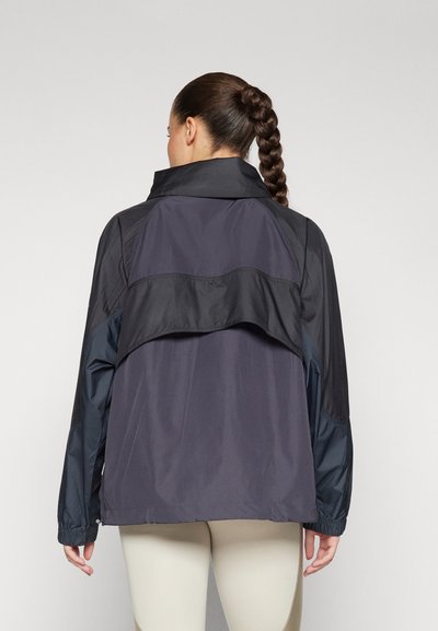 Veste coupe-vent légère de couleur gris foncé avec un col haut, un dos allongé, des poignets élastiques et des détails en panneaux noirs contrastants.
