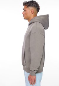 Grå hættetrøje med en frontlomme og ribbede manchetter, der har en justerbar hætte. Båret med lyseblå jeans, vist fra siden.