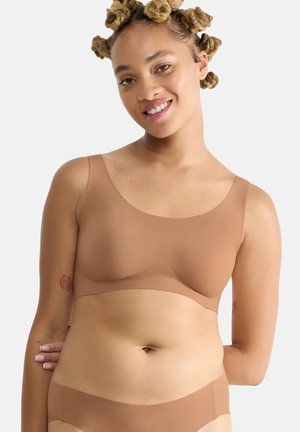 Uśmiechnięta kobieta o jasnobrązowej skórze i blond warkoczach w stylu bantu nosi bezszwowy brązowy biustonosz sportowy oraz dopasowane majtki.