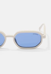 White octagonal sunglasses with blue lenses. Features include a textured frame and "GUESS" logo on the left lens. Lightweight design.