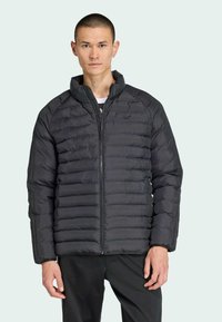 Mladý muž s krátkymi vlasmi v čiernej vypchávanej bunde stojí na svetlom pozadí, ruky má uvoľnené pri svojich stranách.