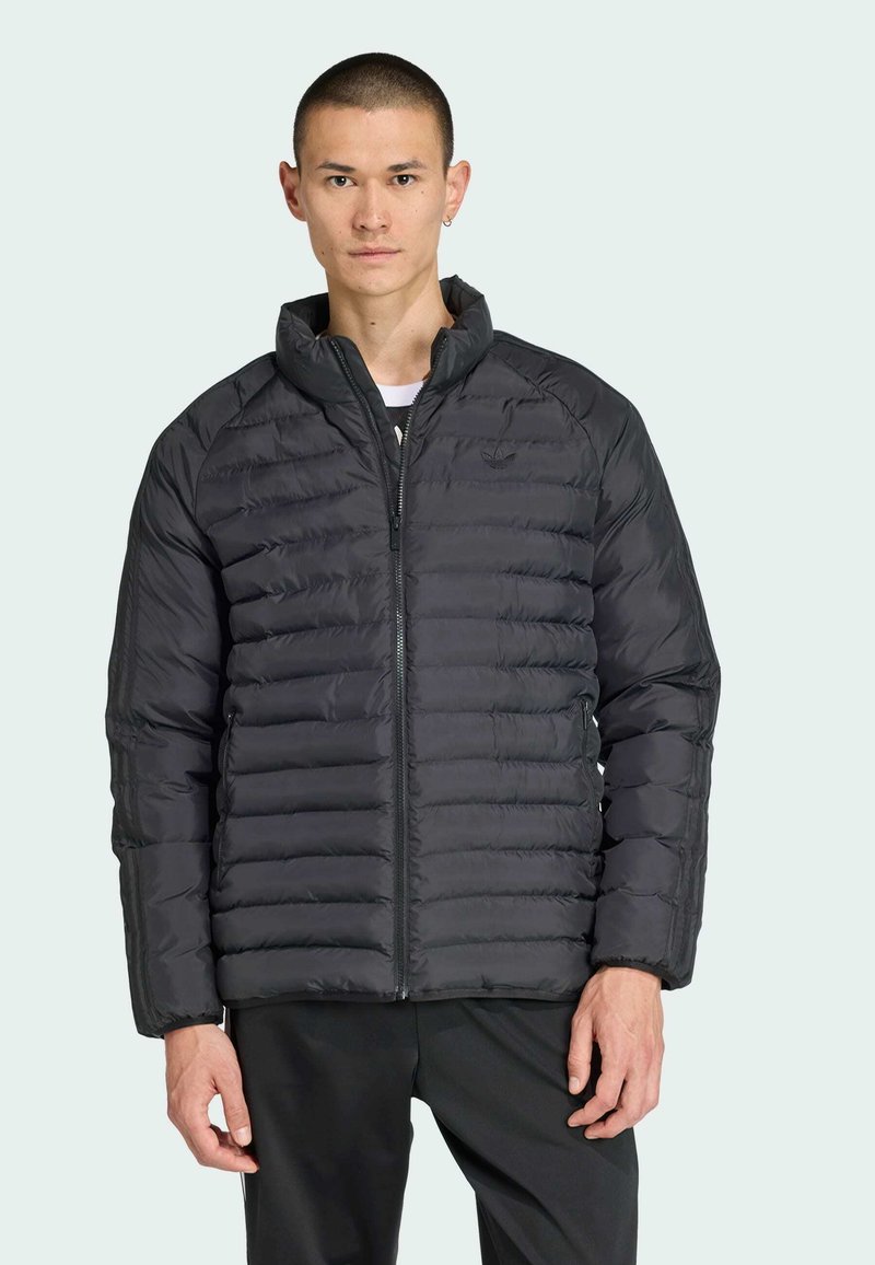 Mladý muž s krátkymi vlasmi v čiernej vypchávanej bunde stojí na svetlom pozadí, ruky má uvoľnené pri svojich stranách.