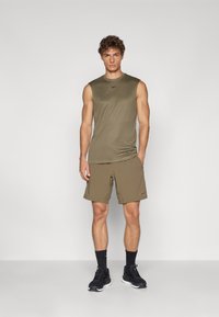 Ελαιόχρωμο αμάνικο τοπ και ταιριαστά σορτς, φτιαγμένα από ελαφρύ υλικό. Μαύρα αθλητικά παπούτσια με υφασμάτινα στοιχεία και σκούρες κάλτσες.