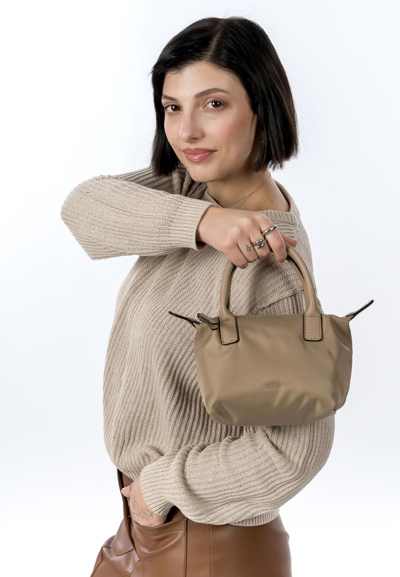 Kompakt beige håndtaske med en glat tekstur, dobbelte korte håndtag og sorte accenter. Modellen har en lys sweater og brune bukser på.