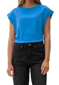 Kobieta w jasnoniebieskiej bluzce bez rękawów, włożonej w czarne dżinsy z wysokim stanem, stoi z relaksującymi ramionami wzdłuż ciała.