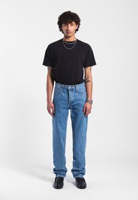 Černé tričko, světle modré rovné džíny a černé boty; model s kudrnatými vlasy stojí na pozadí s bílou plochou.