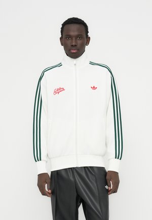 White zip-up track jacket featuring green stripes along the sleeves and red embroidered logos. Worn with black leather pants.