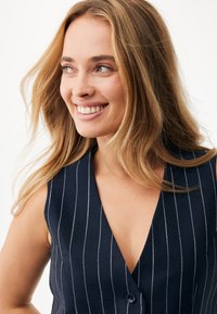 Granatowy, bezrękawnik z pionowymi białymi cienkimi paskami, dekoltem w kształcie litery V i pojedynczym guzikiem z przodu. Gładka faktura tkaniny.