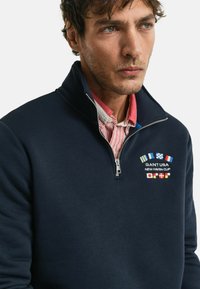 Felpa con zip blu navy e colletto alto, con ricami di bandiere colorate e testo. Realizzata in un tessuto morbido, con uno strato interno strutturato.