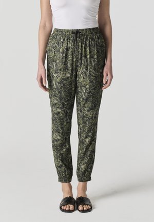 Mujer con pantalones jogger verdes con estampado floral, tobillos elásticos y cordón, camiseta sin mangas blanca y sandalias negras tipo chancla.