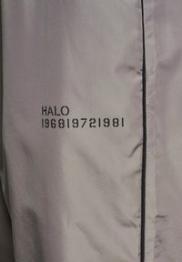 Ljusgrå jacka i material med slät struktur; har svart tryckt text "HALO 196819721981" och en kontrasterande dragkedjedetalj.