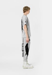 La camiseta de manga corta gris y los pantalones de chándal a juego presentan gráficos de alas negras. Completado con botas blancas, el conjunto tiene una estética moderna y deportiva.