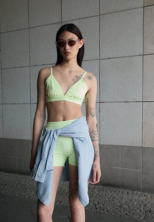 Neon zelený bikiny top a šortky doplněné světle modrou texturovanou košilí uvázanou v pase. Kolo sluneční brýle završují vzhled.
