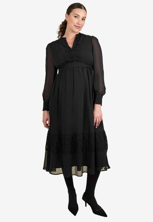 Musthave mustrõngastega must raseduskleit, millel on pikad läbipaistvad varrukad, pitsaktsentidega rinnas ja ääres ning voolav seeliku disain. Selga on pandud kontsad koos mustade sukkpükstega.