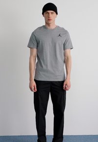 Tricou gri din bumbac cu un logo negru Air Jordan, asociat cu pantaloni cargo negri. Modelul poartă o căciulă tricotată neagră. Fundal minimalist.