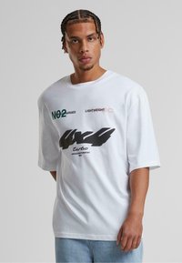 Bijela oversized majica od pamuka, sa crnim i zelenim grafičkim natpisima, uključujući "NO2," "LIGHTWEIGHT," i "turbo," te okruglim izrezom.