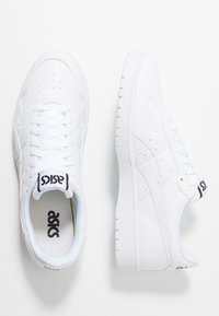 Bílá teniska s texturovaným koženým svrchním dílem, perforovanou špičkou, plochými tkaničkami a polstrovaným límcem. Na jazyku a boku je viditelné černé logo.