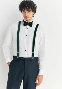 White dress shirt with black buttons, green velvet bow tie, and green suspenders paired with dark trousers. Smooth textures and tailored fit.