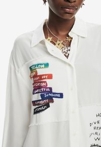 Camicia bianca con bottoni, caratterizzata da strisce di stoffa con etichette testuali colorate. Il tessuto ha una texture leggermente sfrangiata e un design del colletto semplice.