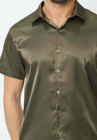 Chemise à manches courtes, de couleur olive, fabriquée en tissu lisse et brillant, avec un col et six boutons sur le devant.