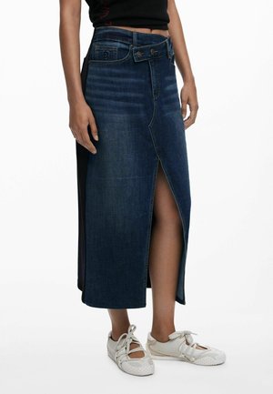 Femme portant une jupe midi en jean bleu foncé avec une fente devant et des baskets blanches à lacets, debout sur un fond blanc.