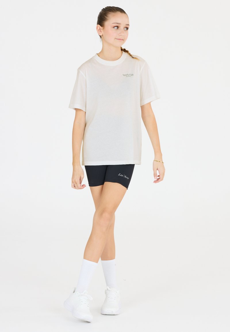 T-shirt blanc en coton à manches courtes avec un logo sur la poitrine, associé à un short de vélo noir et des chaussures de sport blanches avec des semelles texturées.