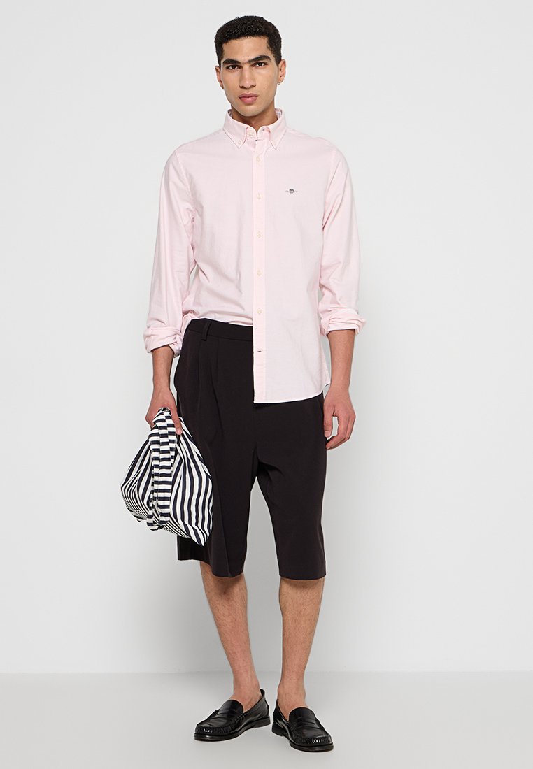 Ein Mann steht selbstbewusst in einem hellrosa Hemd mit Knopfleiste und schwarzen Shorts und hält ein gestreiftes Hemd in der Hand. Er trägt schwarze Slipper.