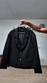 Blazer preto pendurado em um cabide, apresentando lapela com recorte, dois bolsos frontais, fechamento com um único botão e um tecido liso e texturizado.