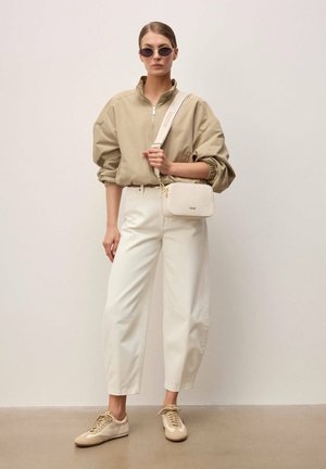 Žena v béžové bundě na zip, bílých kalhotách s vysokým pasem, béžových teniskách, slunečních brýlích a s malou bílou crossbody kabelkou.